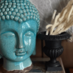 A turquoise ceramic Buddha head figurine is displayed next to a black ornate vase with dried white flowers.